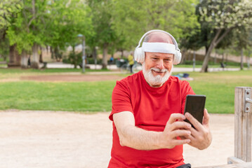 Fototapeta premium Happy sporty Senior man in headband, red shirt and headphones using smartphone in park after workout