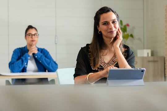 Diverse female colleagues sitting at meeting room table reviewing papers and using tablet computer