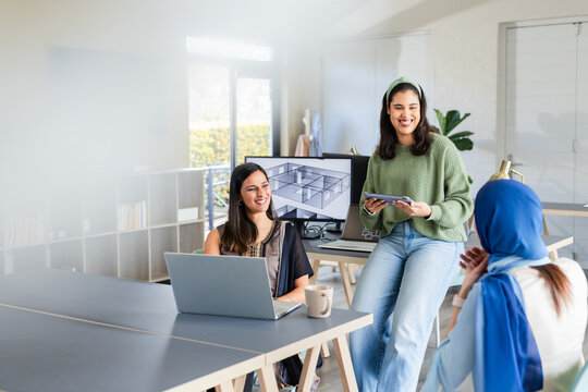 Diverse female coworkers collaborating around workstations in office with laptop tablet and monitor