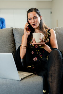 Indian woman sitting at home holding mug pressing smartphone beside silver laptop on gray couch
