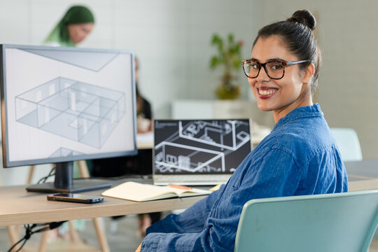 Woman sitting at desk in office working on floor plans on desktop monitor with notebook, smartphone