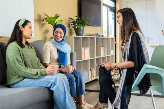 Diverse female colleagues sitting in office lounge holding coffee mugs and using smartphone