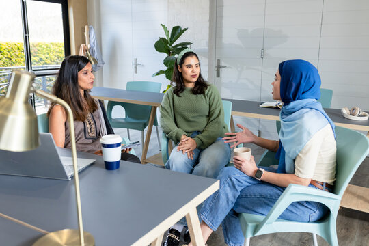 Diverse coworkers sitting around meeting table at break room, sharing laptop and smartphone