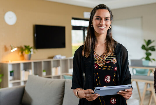 Indian woman standing in modern lounge holding tablet beside mounted flat-screen TV and gray sofa