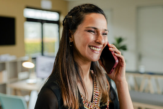 Indian woman wearing kurta holding smartphone to ear while standing by desk in office, copy space