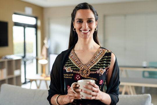 Indian woman standing holding ceramic mug smiling toward camera in living room workspace