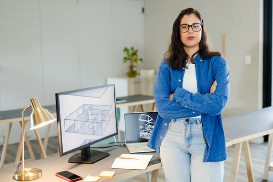 Woman standing arms crossed at worktable in studio reviewing 3D floor plans on monitor and laptop