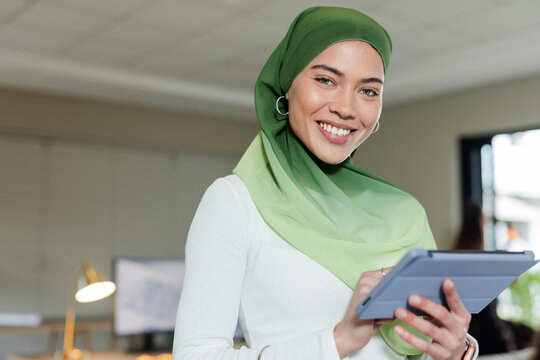 Indian woman wearing hijab standing in open plan office holding tablet by monitors