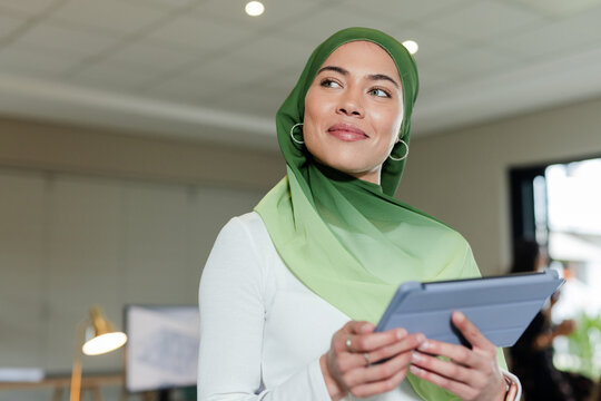 Indian woman standing at meeting room table holding tablet beside brass desk lamp gazing right