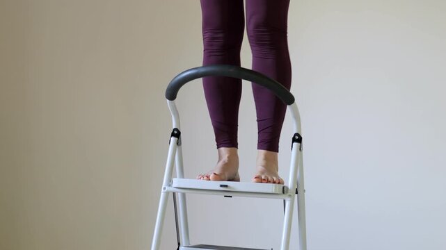 Person is standing on a ladder. The ladder is white and has three steps. The person is wearing black pants