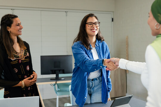 Diverse female coworkers shaking hands around desk in meeting room with laptop, tablet