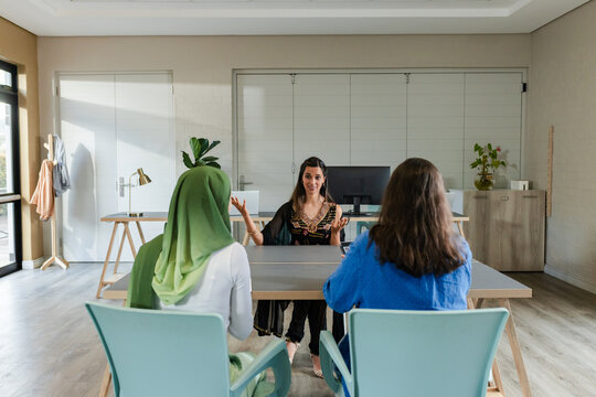 Diverse female coworkers sitting on chairs at table in office reviewing monitor near coat rack