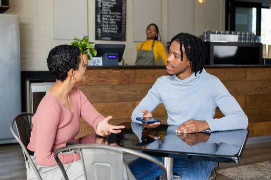 Diverse friends chatting at glossy cafe table using smartphone, barista in apron preparing espresso