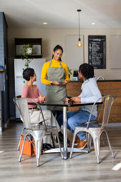 Mid adult woman barista processing digital payment for seated couple at cafe table with chalkboard