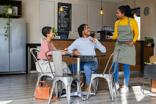 Diverse customers and server interacting at round table in cafe with tablet and chalkboard menu