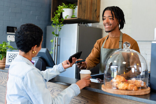African American barista in apron handing card reader to female customer using phone at counter