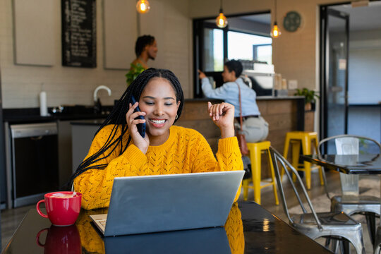Woman wearing mustard sweater holding smartphone and working on laptop at cafe with red mug