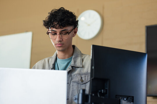 Middle Eastern male student working in classroom with laptop, desktop monitor, whiteboard and clock