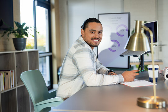 Asian man sitting at modern office workspace holding green pen and smiling beside dual monitors