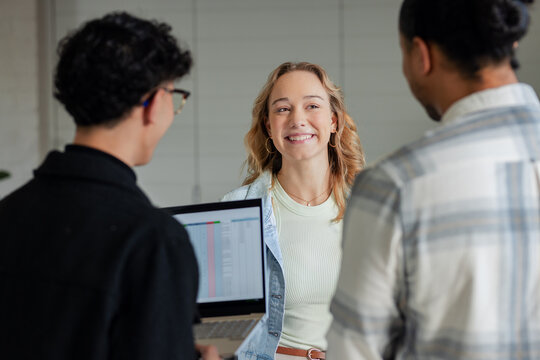 Diverse coworkers standing in office wearing attire smiling and discussing spreadsheet on laptop