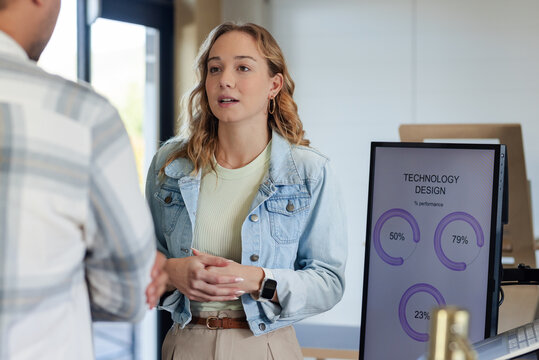 Diverse coworkers speaking and reviewing performance charts on monitor with prototypes in office
