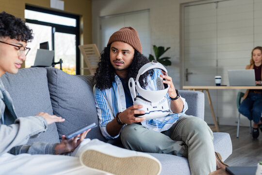 Diverse coworkers holding white futuristic helmet and reviewing specs on tablet in office lounge