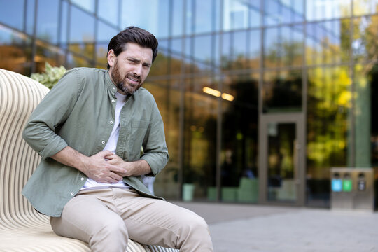 Man experiencing acute stomach pain and discomfort, holding his abdomen with a grimacing expression while sitting on a bench outside a modern building, representing a health issue