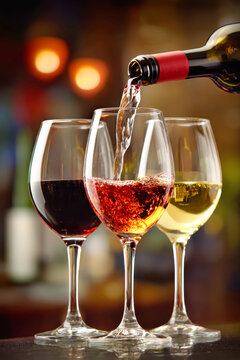 Red wine is being poured into a glass while two other glasses of white and ros? sit nearby against a blurred restaurant background