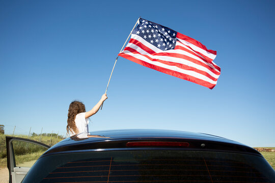 Girl holding American flag out of a car under blue sky
