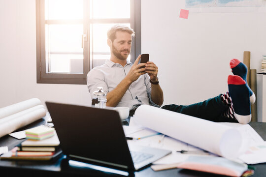Portrait of smiling architect sitting with feet up at desk using cell phone