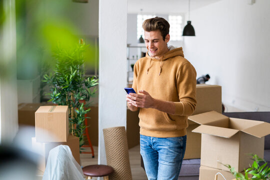 Smiling young man using mobile phone in messy living room during relocation
