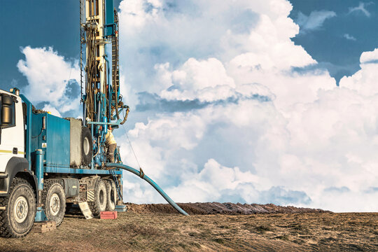 A drilling rig operates in an open area as workers handle equipment and prepare for drilling activities. The sky shows clouds in the background