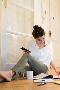 Young woman sitting on the floor with coffee mug and smartphone looking at booklet