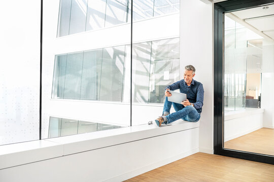Casual businessman sitting on windowsill in office building, using laptp