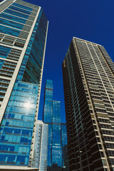 Fototapeta premium Tall buildings reach upward in a city skyline during midday with a clear blue sky and sunlight reflecting off glass windows