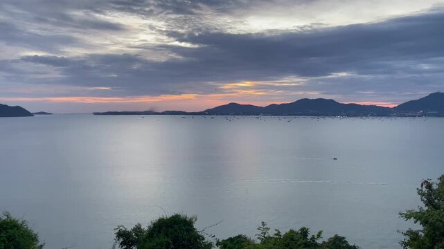 Wide panoramic view of a calm sea at sunset with dramatic clouds, distant islands, and the Phuket coastline silhouetted on the horizon, Thailand