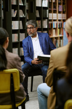 Middle aged South Asian man holding book presenting to group of adults in library setting, engaging audience with focused expression, bookshelves filled with books in background