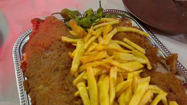Close-up view of traditional Spanish cachopo, a breaded veal fillet stuffed with ham and cheese, served with golden French fries and Padr&oacute;n peppers on a metal tray
