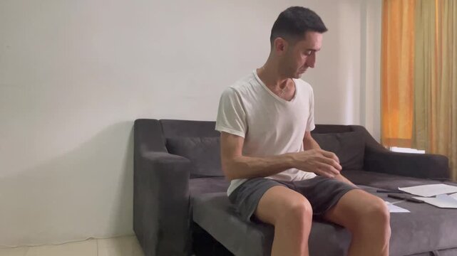 Adult man sitting on a sofa at home tearing paper by hand, expressing frustration or decision-making stress in a simple indoor setting with neutral background