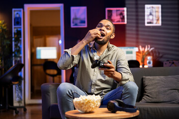 Male gamer eating popcorn while he plays online shooting tournament, having fun with video games in...