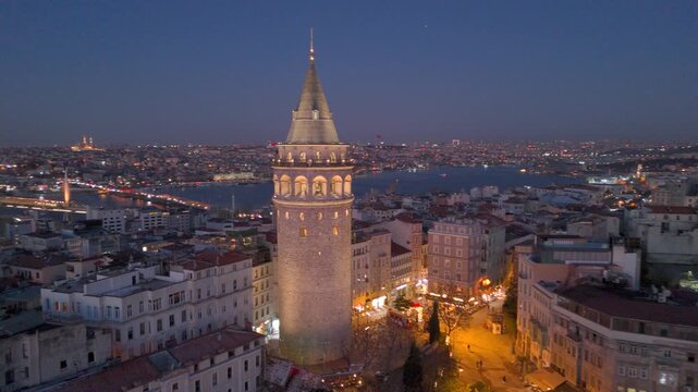 Morning view of Galata Tower illuminated against historic Golden Horn and cityscape in Istanbul with lights illuminating streets and buildings at sunrise. Flying around iconic Galata Tower in sunset.
