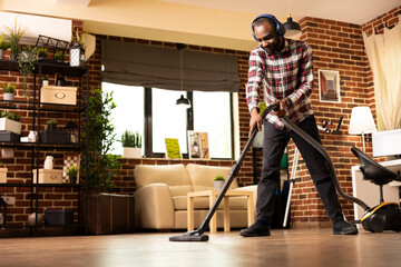 Fototapeta premium Smiling black man with headphones, tidying up apartment while listening to podcast. African american male individual vacuums floors and enjoys music, making weekend cleaning an enjoyable activity.