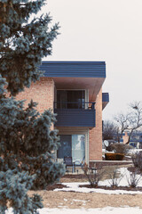 Building in winter with snow on ground and tree in foreground near chairs