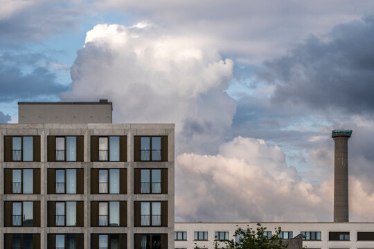 Contemporary urban building skyline with strong facade and window rhythm beside tall chimney under expansive cloud filled sky offering minimal copy space