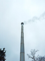 Smoke rises from a tall chimney against a cloudy sky with tree branches visible below the towering structure
