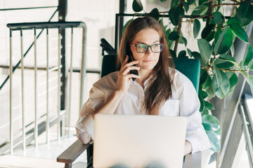 A business woman in a white shirt sits in a chair by the window using a laptop to work.