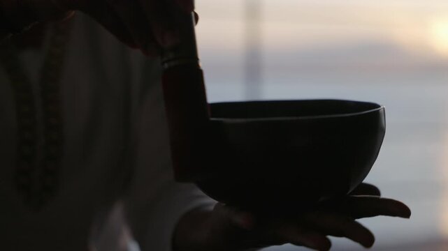 Silhouette of hands with mallet playing a Tibetan singing bowl. Tropical sunrise ocean in the background.