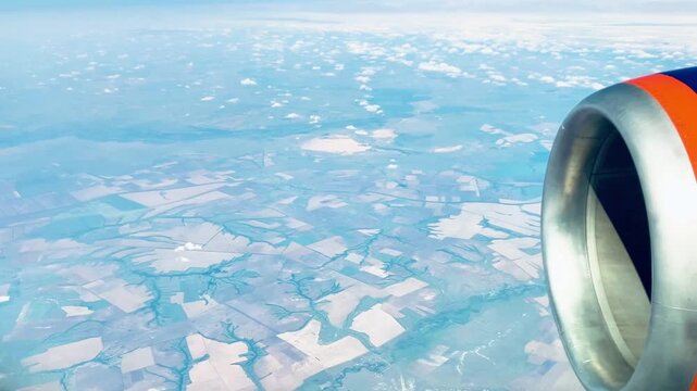 Airplane window view with jet engine flying above clouds and farmland below. Scenic aerial countryside landscape, blue sky. Air travel and transportation concept background