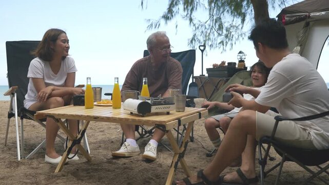 Family camping beach outdoor fun with kids, tent provides shade for relaxed children, multi generational adults enjoy calm trip, sitting at wooden table with drinks, portable stove.
