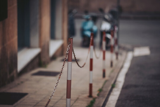 In a serene city street, red and white chain barriers line the sidewalk, directing foot traffic. Parked scooters rest nearby, hinting at the bustle of a new day dawning